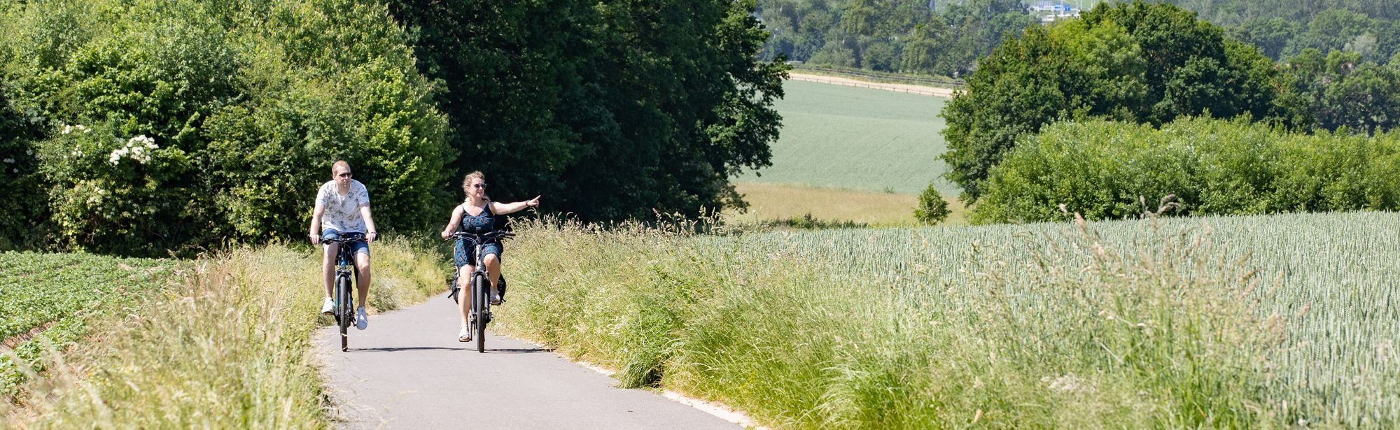 Een koppel fietst door het groene heuvellandschap en de vrouw wijst naar iets in de verte