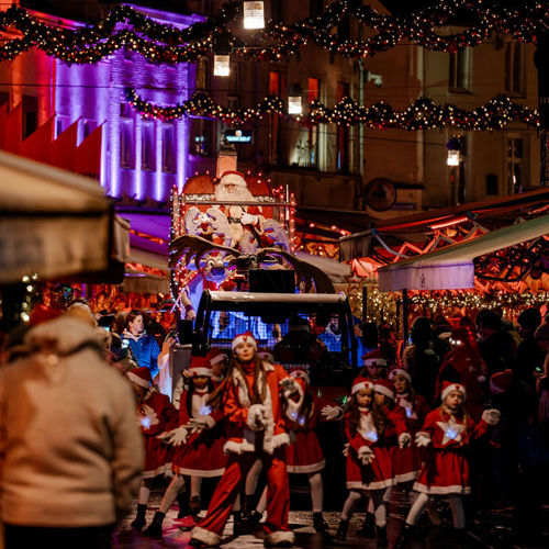 Een dansgroep van meisjes in kerstmannenpakken dansen voor een praalwagen met daarop de Kerstman tijdens Kerststad Valkenburg Parade