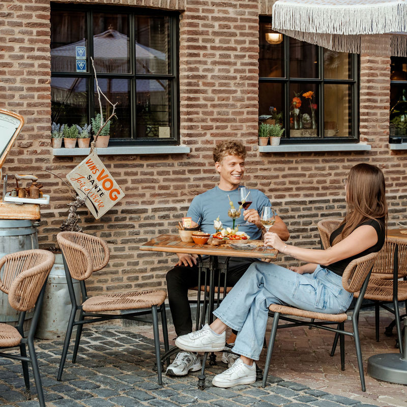 Een man en vrouw proosten met een wijntje bij Wijnterras D'Hunes naast karakteristieke attributen van een oude boerderij. 